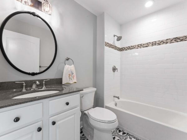 Modern bathroom in Bucky County, PA with light grey walls, white tile and bath, sink with dark grey countertop, and round mirror.