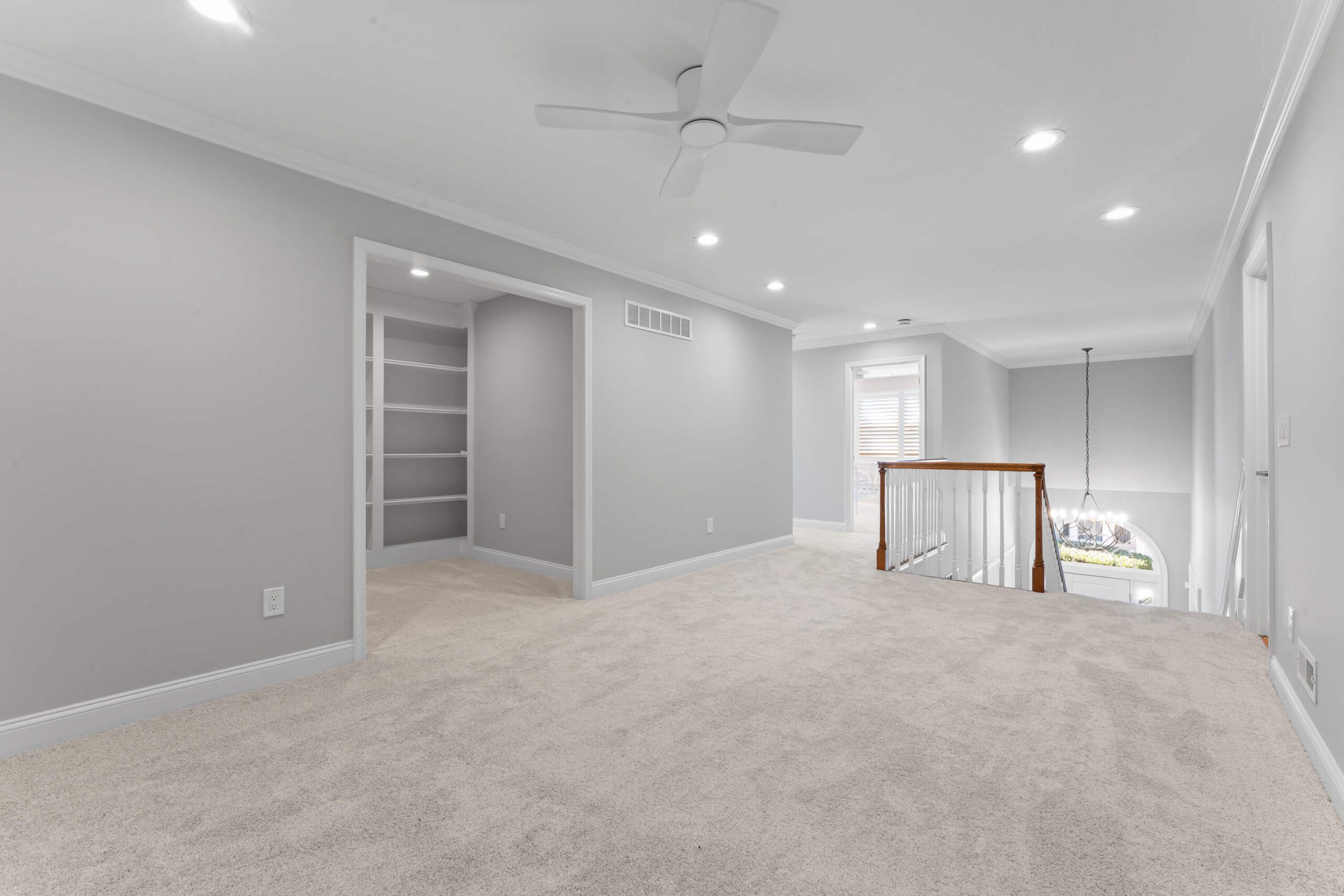 Spacious carpeted hallway with gray walls, recessed lighting, ceiling fan, and open staircase with wooden railing.