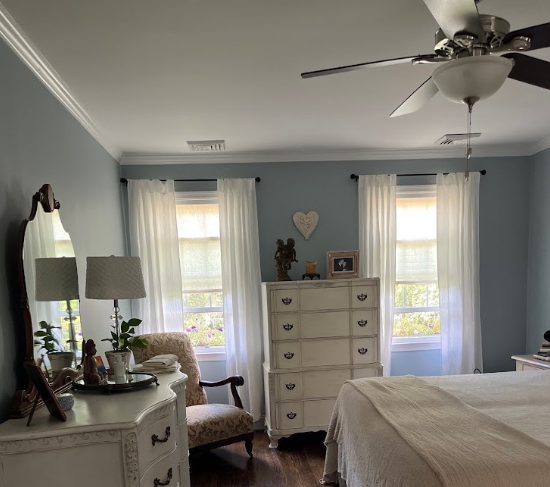 Bedroom with light blue walls, white curtains, ceiling fan, dresser, and armchair, creating a cozy, elegant atmosphere.
